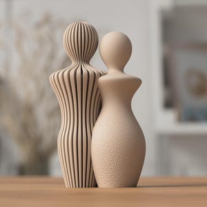 May include: Two beige decorative sculptures. One is a solid form, the other is made of vertical wooden slats. The sculptures are placed close together, suggesting a couple or pair. The background is blurred, focusing attention on the objects.