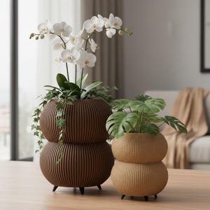 May include: Two decorative, stacked, spherical planters. One is a dark brown colour and holds a white orchid. The other is a light brown colour and holds a green plant. Both planters have a vertical, ribbed design and sit on small black legs.
