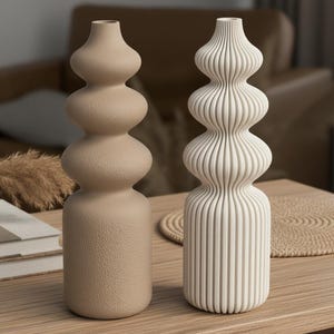 Könnte beinhalten: Zwei dekorative Vasen auf einem Holztisch. Eine Vase ist beige mit strukturierter Oberfläche und bauchiger Form. Die andere ist weiß mit vertikalen Rippendetails. Die Vasen stehen neben einer kleinen Schale und Büchern.