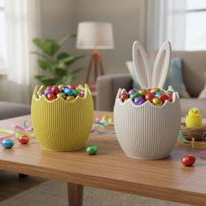 May include: Two decorative Easter egg-shaped containers filled with colourful foil-wrapped chocolate eggs. One container is yellow, the other is grey with white bunny ears. The containers sit on a wooden table with decorative ribbons.