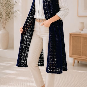 May include: A long, sleeveless, navy blue crochet vest featuring an open-knit pattern. The vest is worn over a white button-down shirt with thin vertical stripes, paired with beige trousers, a tan belt, and beige loafers.