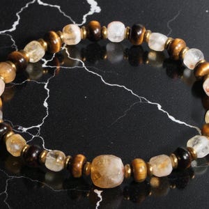 May include: A beaded bracelet featuring a mix of brown, gold, and clear gemstones. The bracelet has a variety of bead shapes, including round and rectangular, strung together with gold-colored spacers. The bracelet is displayed on a black surface.