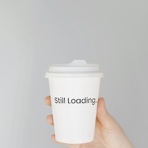 May include: A white paper coffee cup with a lid, held in a hand. The cup has the words "Still Loading..." printed in black. The background is a solid, light gray color.