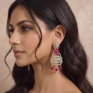 以下が含まれることがあります： ゴールドトーンの優雅なイヤリング。上部に花のモチーフ、バゲットカットのクリスタルが連なるデザイン。ピンク色の宝石と、3つのしずく型のピンク色の宝石が特徴です。