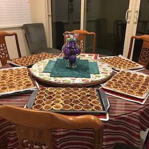Puede incluir: Una mesa redonda preparada para una reunión, cubierta con un mantel a rayas rojas y blancas. Varias bandejas de mini tartas de nueces pecanas están dispuestas sobre la mesa, junto con un jarrón decorativo lleno de adornos morados. Sillas de madera rodean la mesa.