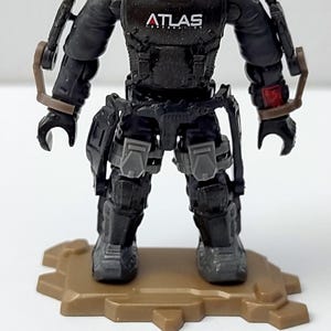 May include: A small plastic action figure wearing a black exoskeleton suit with the word ATLAS printed on the chest, a dark beanie, and grey leg armour, standing on a tan base with a separate black and grey toy rifle placed in front.