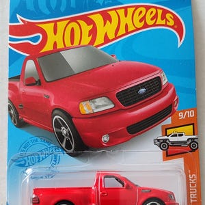 May include: A red Hot Wheels die-cast model of a '99 Ford F-150 SVT Lightning pickup truck. The toy car is on a blue and orange card with the Hot Wheels logo and the text "HW Hot Trucks". The model is 9/10 in the series.