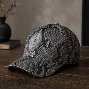 Op de afbeelding: Een baseballpet met een gestructureerd slangenleerpatroon in grijs en zwart. De pet heeft een gestructureerde kroon, een gebogen klep en een kleine knoop aan de bovenkant. Het materiaal vertoont een gedetailleerde print met schubben.