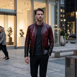 Può includere: Giacca in pelle bordeaux con texture a pelle di coccodrillo. La giacca ha colletto e polsini neri. La persona indossa una maglietta grigia e pantaloni neri. Lo sfondo mostra una strada cittadina con edifici e vetrine.