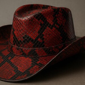 Op de afbeelding: Een cowboyhoed met een dieprood en zwart slangenleerpatroon over het gehele oppervlak. De hoed heeft een gestructureerde kroon met een ingeknepen bovenkant en een brede, gebogen rand, geaccentueerd door een bijpassende band rond de basis.