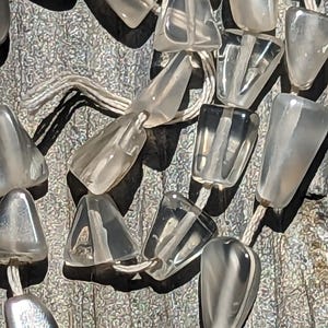 May include: A strand of translucent, faceted, gray gemstone beads strung on a white cord. The beads are shaped like teardrops and are arranged in a random pattern.