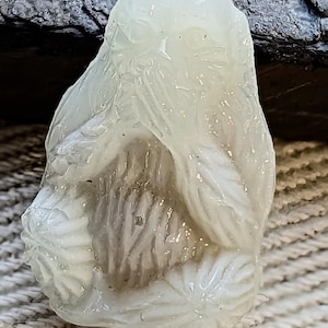 May include: A white, translucent, carved glass pendant with a detailed floral design. The pendant has a smooth, polished surface and a slightly irregular shape.