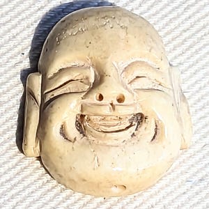 May include: A carved ivory pendant featuring a smiling Buddha face with closed eyes. The pendant is a light cream color and has a hole drilled through the bottom for hanging.