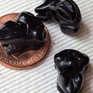 May include: Three small, glossy black frog figurines. The frogs are carved with detailed features and sit on a white surface next to a penny. The frogs are a dark, solid color.