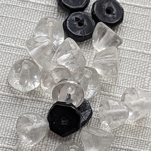 Puede incluir: Una colección de cuentas de vidrio transparente facetadas y cuentas negras en forma de botón. Las cuentas transparentes tienen una apariencia cristalina y brillante, mientras que las cuentas negras son opacas y tienen una textura suave. Las cuentas están dispersas sobre una superficie clara y texturizada.