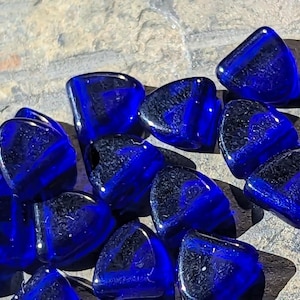 May include: A collection of deep blue glass beads with a textured, swirled interior. The beads are irregularly shaped, with rounded edges and a glossy finish. The beads are scattered on a rough, light-colored surface, creating a contrast of textures and colors.
