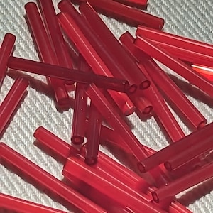 Op de afbeelding: Een close-up van talrijke doorschijnende rode glazen buisjes. De buisjes zijn cilindrisch met open uiteinden en liggen verspreid op een getextureerd oppervlak. De belichting benadrukt de glanzende textuur en de levendige rode kleur van het glas.