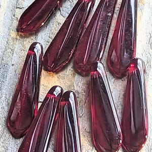 Könnte beinhalten: Eine Ansammlung tropfenförmiger Perlen in tiefem Burgunderrot. Jede Perle hat eine abgerundete Oberseite mit einem kleinen Loch zum Auffädeln und eine spitz zulaufende Unterseite. Die Perlen liegen auf einer strukturierten, hellgrauen Oberfläche.