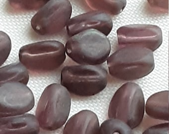 May include: A selection of oval, translucent beads in shades of purple and mauve. The beads have a smooth, polished look and are scattered on a white surface. These beads are suitable for jewellery making and craft projects.
