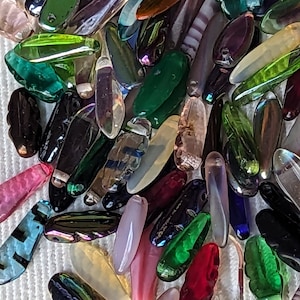 May include: A collection of colorful glass beads in various shapes and sizes. The beads are mostly oval and have a smooth, glossy finish. The colors include green, blue, red, pink, purple, and clear.