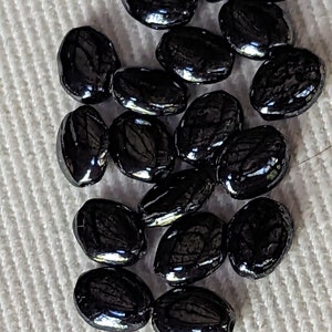 May include: A collection of black oval-shaped beads with a glossy finish. The beads are scattered on a white textured surface.