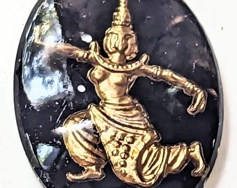 May include: An oval-shaped, dark background with a gold-colored, ornate figure in a dynamic dance pose. The figure is wearing a detailed headdress and traditional clothing, with arms outstretched.