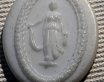 May include: White cameo with a raised design of a woman in a flowing robe holding a staff. The cameo is oval shaped and has a rope-like border.