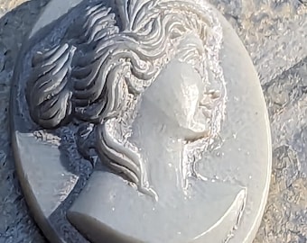 May include: A gray stone cameo featuring a detailed profile of a woman with her hair pulled back. The cameo is oval shaped and has a smooth, polished surface.