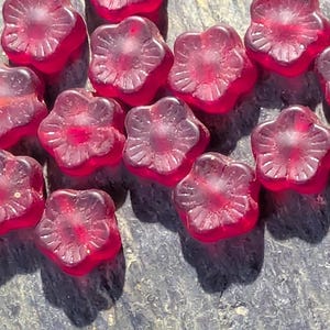 Puede incluir: Un conjunto de cuentas translúcidas en forma de flor, en tonos rojos. Las cuentas tienen un diseño floral detallado y proyectan sombras sobre una superficie gris texturizada. El tema general es la fabricación de joyas.