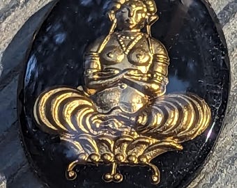 May include: An oval-shaped, black stone pendant with a gold-colored, seated Buddha figure. The Buddha is in a meditative pose, with hands resting in the lap. The figure has detailed features, including a headdress and ornate clothing. The pendant is approximately 2 inches in length.