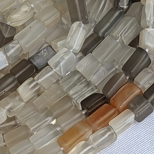 May include: A strand of translucent, square-shaped beads in shades of gray, brown, and orange. The beads are strung together on a clear thread.