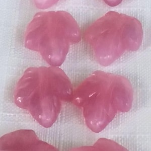 May include: A collection of pink, leaf-shaped beads. The beads have a translucent quality and are strung together. The color is a soft, rosy pink, and the beads are arranged on a white surface. These beads could be used for jewelry making.