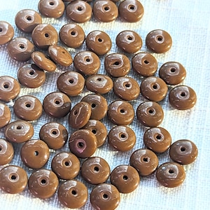 Puede incluir: Un conjunto de pequeñas cuentas redondas marrones, con forma de rosquilla y un agujero central. Las cuentas tienen un acabado brillante y están dispersas sobre una superficie blanca. Adecuadas para la creación de joyas y proyectos de artesanía.