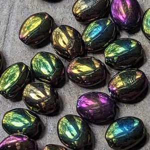 May include: A collection of iridescent oval-shaped glass beads in shades of green, blue, purple, and gold. The beads have a metallic sheen and are scattered on a textured surface.