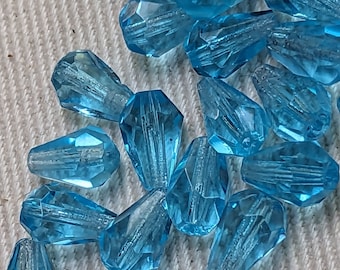 May include: A collection of teal blue faceted glass teardrop beads. The beads are scattered on a white textured surface.