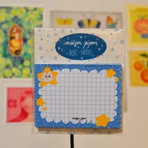 Peut inclure: Un bloc-notes avec un design bleu et blanc, avec un motif quadrillé et des illustrations d'étoiles. Le bloc-notes porte les inscriptions "maison pipon" et "BLOC-NOTES" dans une police fantaisiste. Le bloc-notes est dans une pochette en plastique transparent.