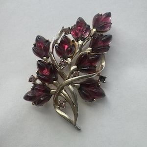 May include: Vintage brooch featuring a floral design. The brooch has a gold-tone metal base with a cluster of deep red, faceted glass leaves. Small, round, purple-toned rhinestones are scattered throughout the design, adding sparkle.