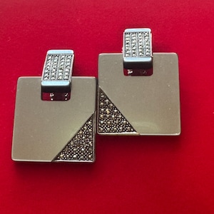 May include: A pair of silver-toned square earrings with a geometric design. Each earring features a rectangular top with a small, rectangular section covered in small, sparkling stones. A triangular section on each earring is also covered in the same stones.