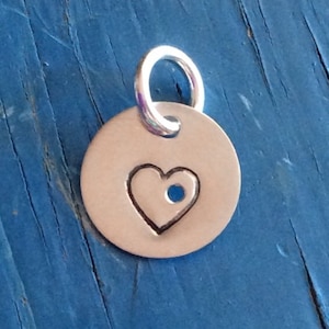 May include: A silver charm with a heart-shaped cutout and a small hole in the center. The charm is attached to a silver loop.