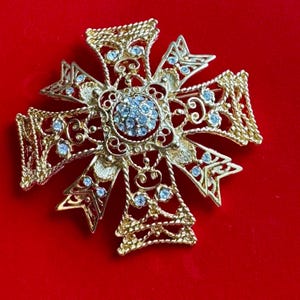 May include: A gold-toned cross-shaped brooch with intricate filigree and rope-like detailing. The center and arms of the cross are adorned with small, sparkling blue gemstones. The brooch is set against a red background.