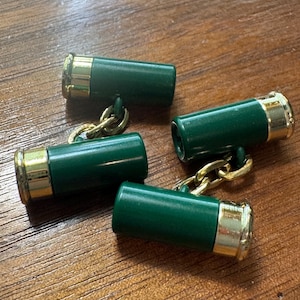 May include: A pair of cufflinks designed to resemble shotgun shells, featuring dark green cylindrical bodies with polished gold-tone metal bases and gold-tone chain links connecting the two ends of each cufflink for a secure closure.