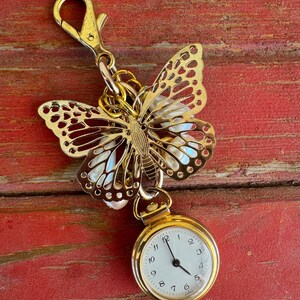 May include: Gold-tone butterfly charm with a small pocket watch attached. The watch has a white face with black hands and black Roman numeral markings.