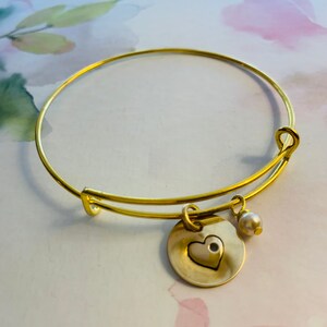 May include: Gold-toned adjustable bangle bracelet. The bracelet features a round charm with a heart cutout and a small pearl charm. The heart charm has a gold outline and a silver center. The bracelet is on a soft, floral background.