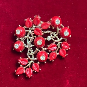 May include: A vintage brooch featuring a floral design with red, round, and leaf-shaped elements. The brooch has a silver-tone metal base and is adorned with clear, round rhinestones. The brooch is set against a red velvet background.