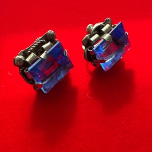 May include: A pair of vintage clip-on earrings. Each earring features a square, faceted, blue gemstone set in a silver-toned metal frame. The frame has decorative rope and floral elements. The earrings are displayed on a red background.