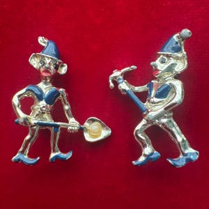 May include: Two vintage clown pins. Each pin features a silver-toned clown figure with blue and red accents. One clown holds a shovel-like object with a pearl-like center, while the other holds a blue instrument.