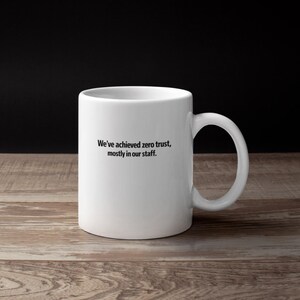 May include: White ceramic mug with a curved handle. The mug has black text that reads, "We've achieved zero trust, mostly in our staff." The mug is on a wooden surface with a black background.