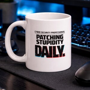 May include: White ceramic mug with the text "CYBER SECURITY PROFESSIONAL PATCHING STUPIDITY DAILY" in black, red, and blue. The mug has a large handle and is sitting on a black surface.