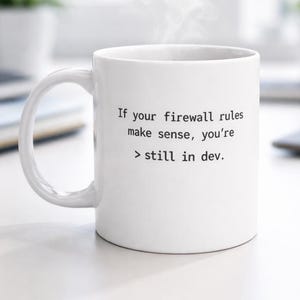 Puede incluir: Taza de café de cerámica blanca con el texto "If your firewall rules make sense, you're > still in dev." La taza tiene un asa curva y está sobre una superficie blanca. El vapor se eleva de la parte superior de la taza.