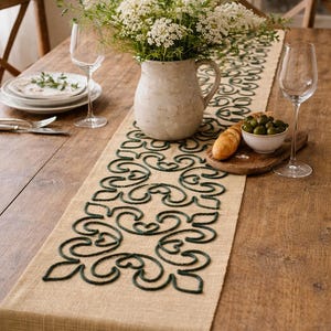 Op de afbeelding: Een rustieke eettafelsetting met een beige tafelloper met een donkergroen geborduurd ontwerp. Een witte keramische kan bevat een boeket witte bloemen. Wijnglazen, borden en een kom olijven maken de scène compleet.